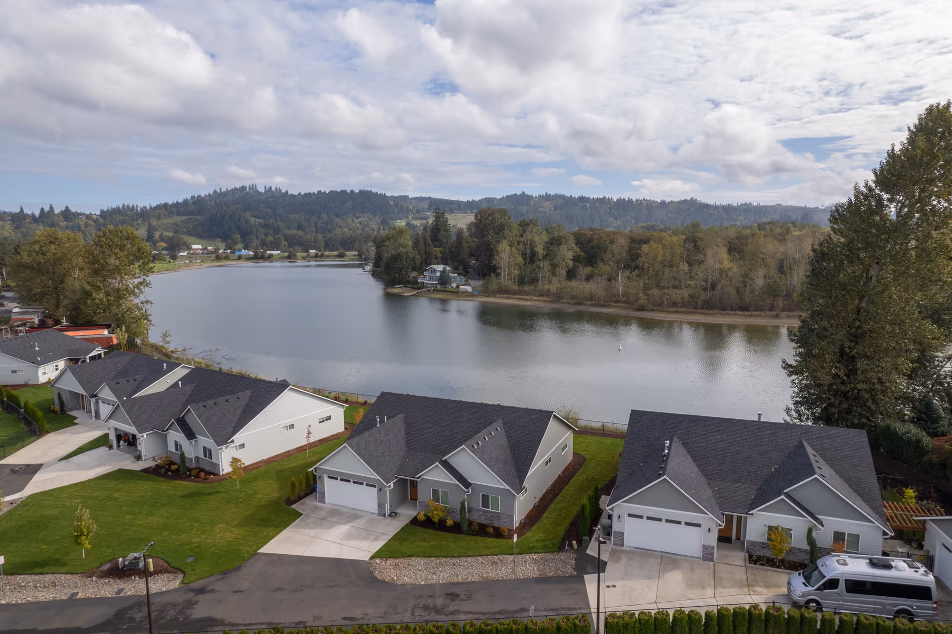 Aerial view of a residential area with several single-story houses with dark roofs and garages, situated near a large body of water with trees and hills in the background under a partly cloudy sky.