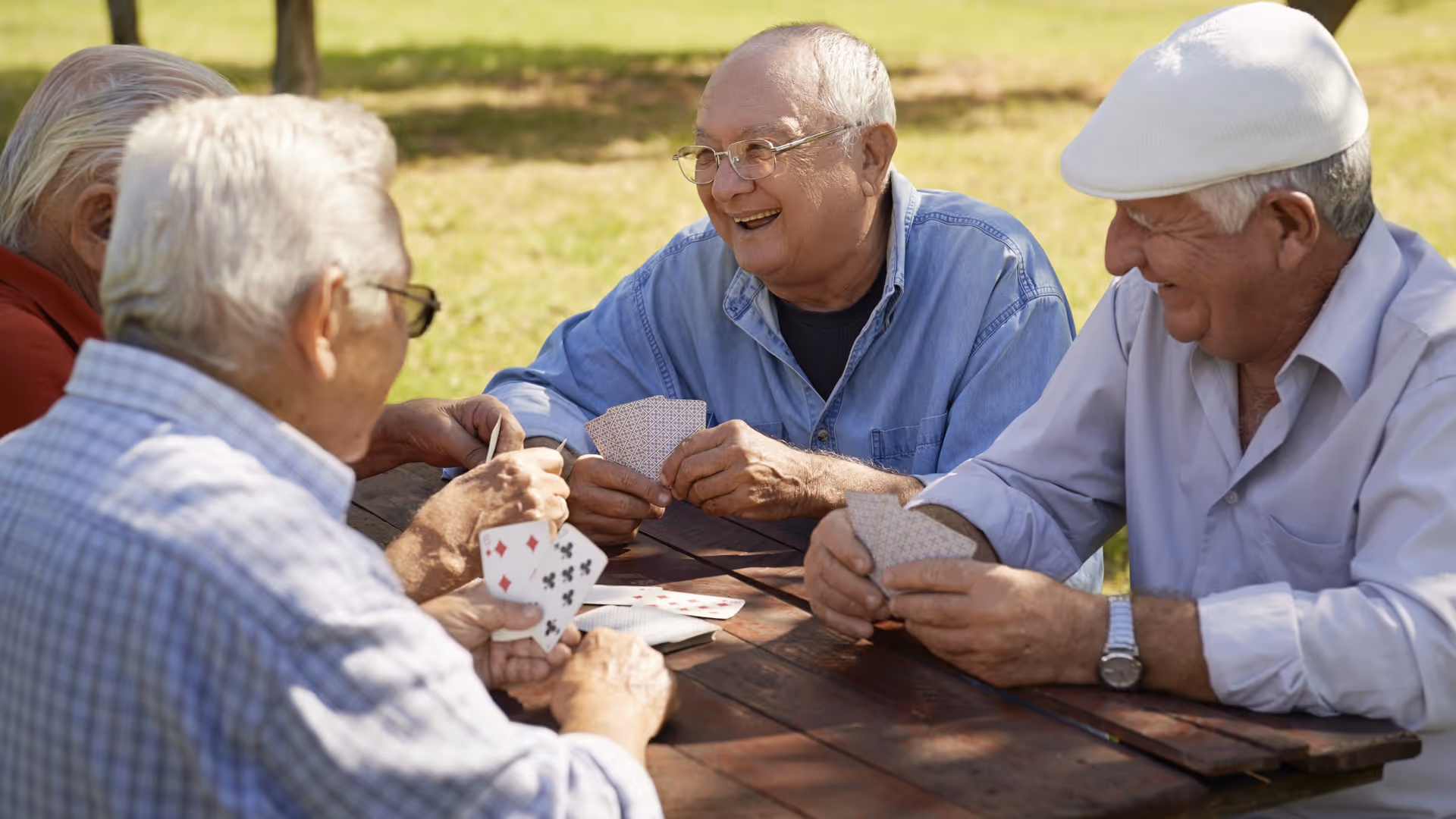 Four elderly men sitting around a wooden picnic table outdoors, playing cards and smiling, with green grass and trees in the background.