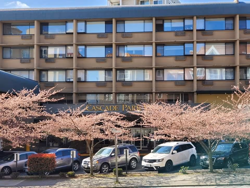 Front exterior view of a multi-story building with large windows and a sign that reads 'Cascade Park'. There are several cars parked in front of the building and pink blossoming trees lining the sidewalk.