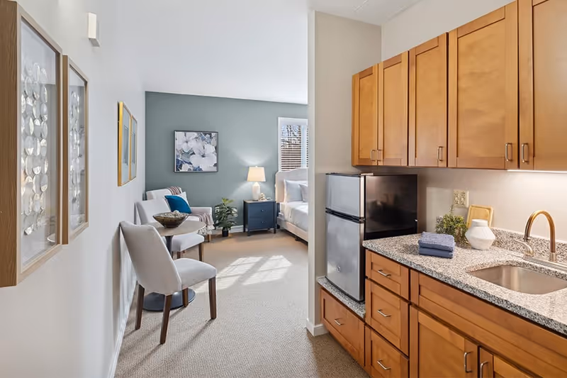 A cozy assisted living apartment featuring a small kitchenette with wooden cabinets, a granite countertop, a sink, and a mini refrigerator. Adjacent to the kitchenette is a sitting area with two upholstered chairs and a small round table. In the background, there is a bedroom area with a bed, a nightstand with a lamp, a plant, and a window with blinds. The walls are decorated with framed artwork, and the room is well-lit with natural light.