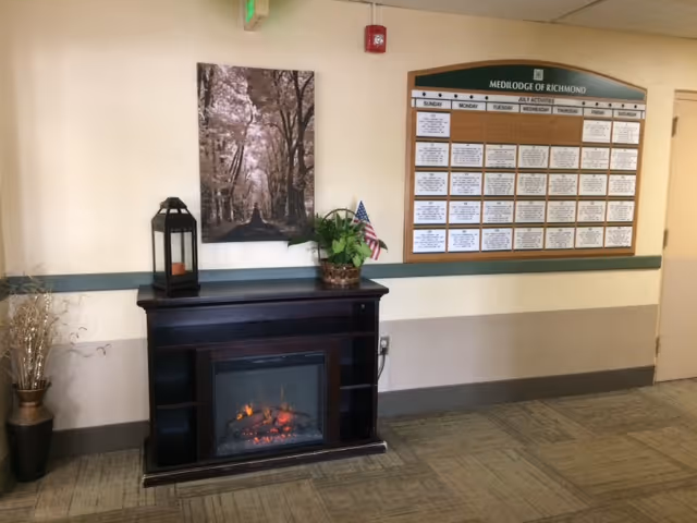 Interior view of a hallway or common area in Medilodge of Richmond featuring a dark wooden electric fireplace with a lantern and a potted plant on top. Above the fireplace is a framed picture of a tree-lined path. To the right, there is a large activity schedule board labeled Medilodge of Richmond with days of the week and various activities listed. The walls are painted beige with a green trim, and the floor is carpeted.