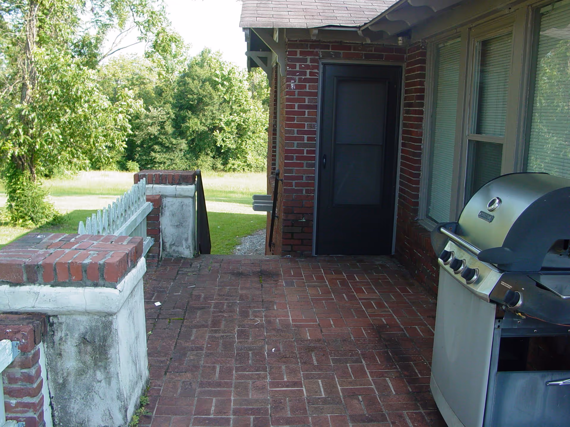 Brick front porch with a stainless steel gas grill, a screen door, and a grassy yard with trees beyond.