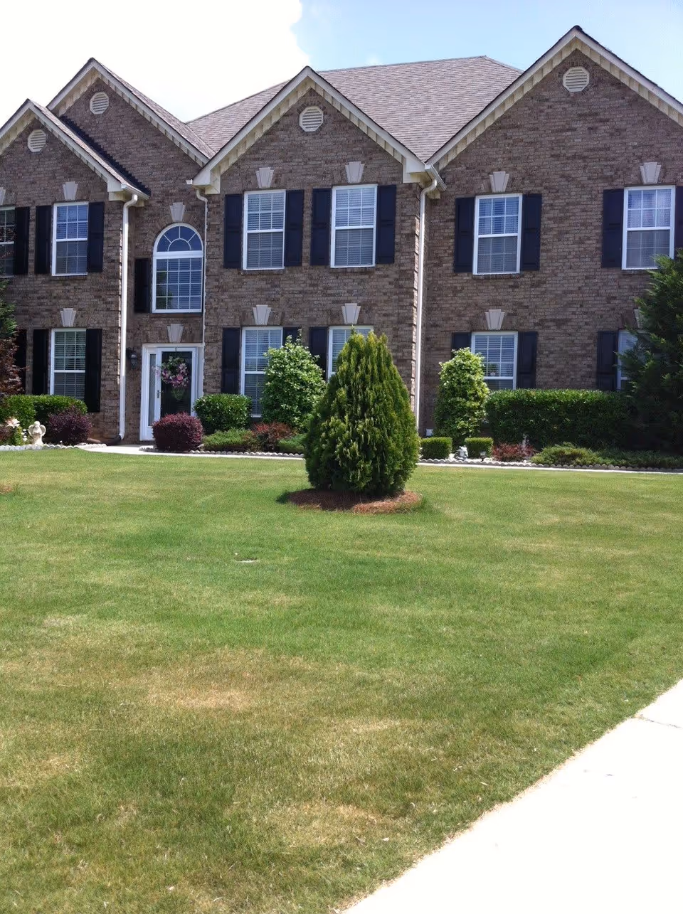Front exterior view of a two-story brick building with multiple windows featuring black shutters. The building is surrounded by a well-maintained lawn with various shrubs and small trees, and a clear blue sky is visible above.