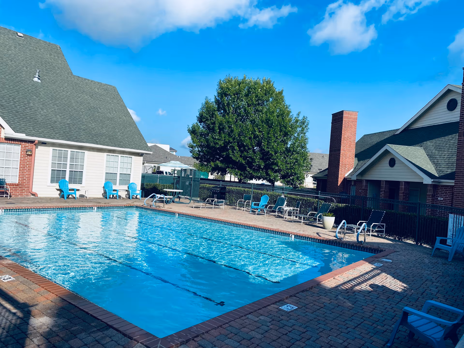 Outdoor swimming pool surrounded by lounge chairs and brick residential buildings under a clear blue sky.