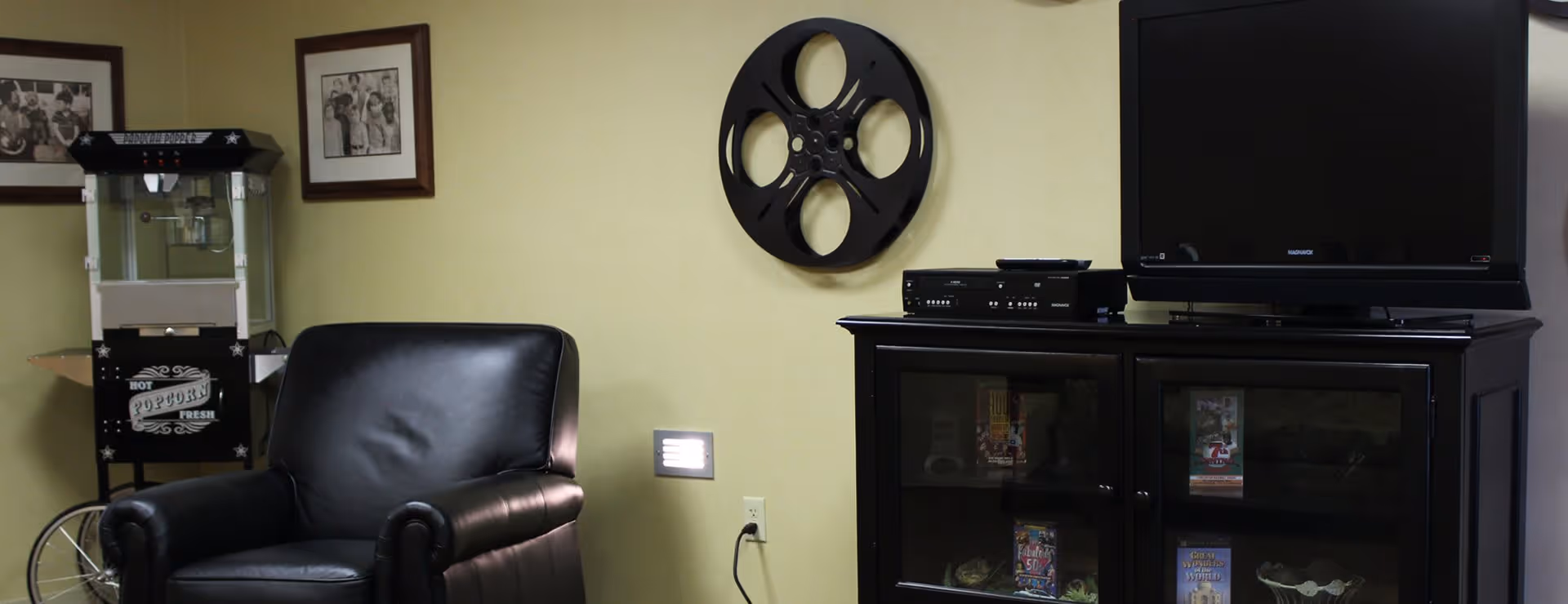A cozy entertainment area with a black leather armchair, a popcorn machine on a stand, a wall-mounted decorative film reel, and a black cabinet holding a flat-screen TV and media devices. The cabinet has glass doors displaying DVDs and decorative items inside.