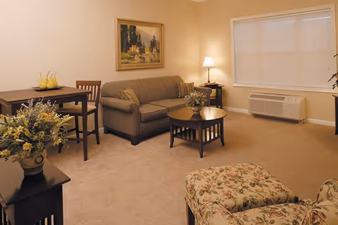 A cozy living room with a beige carpet, a gray sofa with cushions, a wooden coffee table with a floral arrangement, a small dining table with two chairs, a floral upholstered armchair, a side table with a lamp, a large window with blinds, and a wall painting.