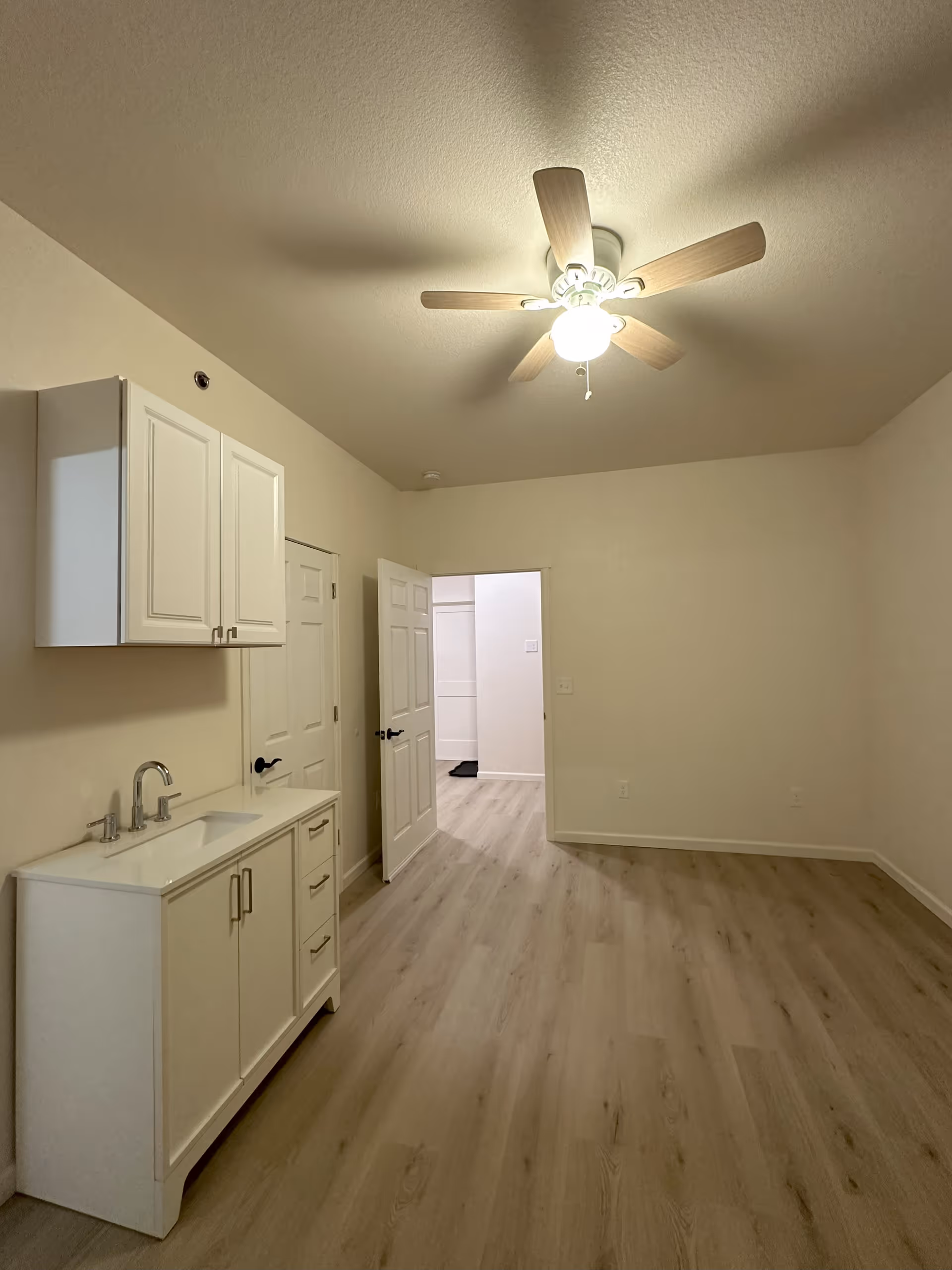 Empty room with light wood flooring, beige walls, a white ceiling fan with light, a white cabinet with a sink and faucet on the left wall, and an open door leading to another room.