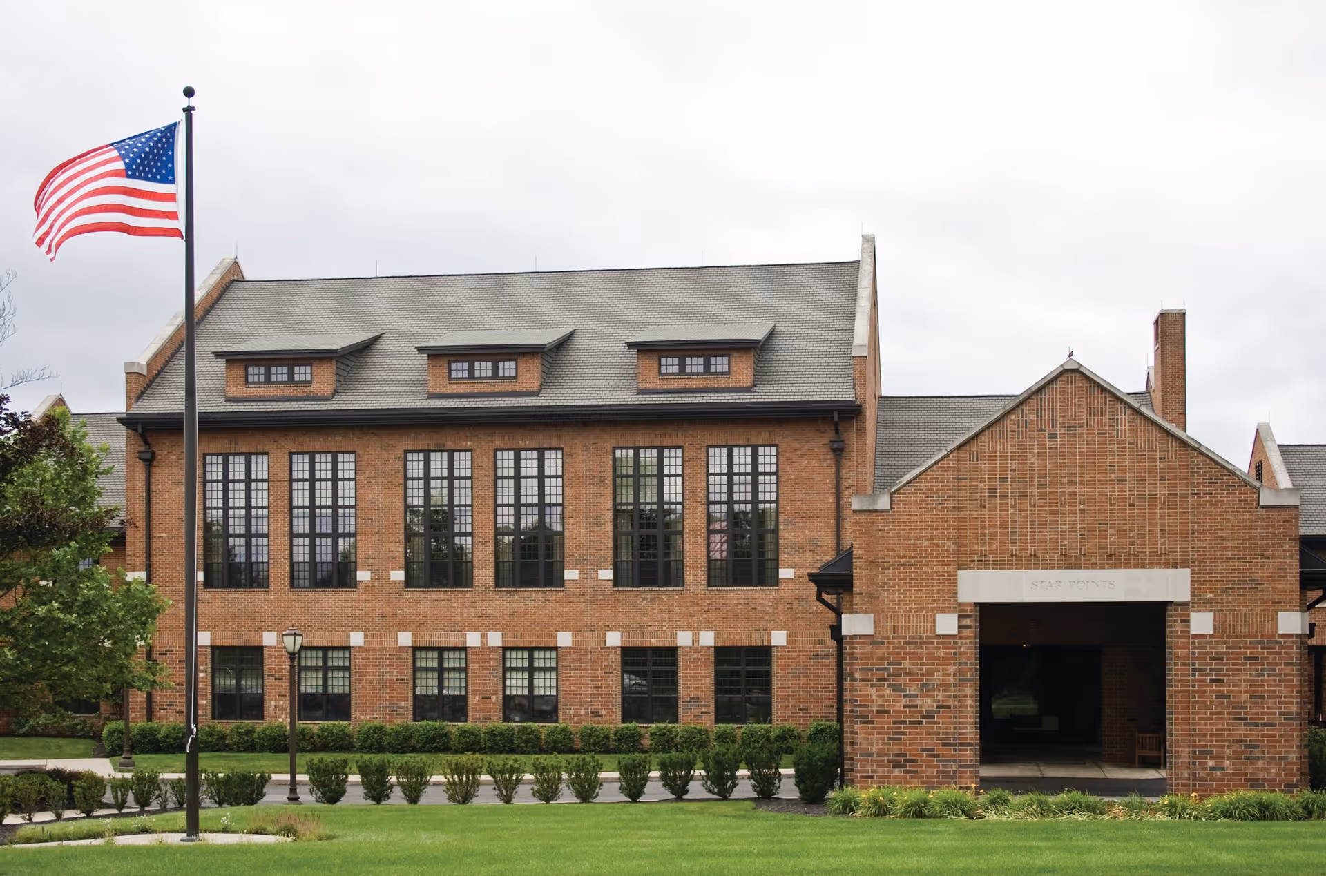 Front exterior of a large brick building with tall windows, an American flag on a flagpole, and a manicured lawn.