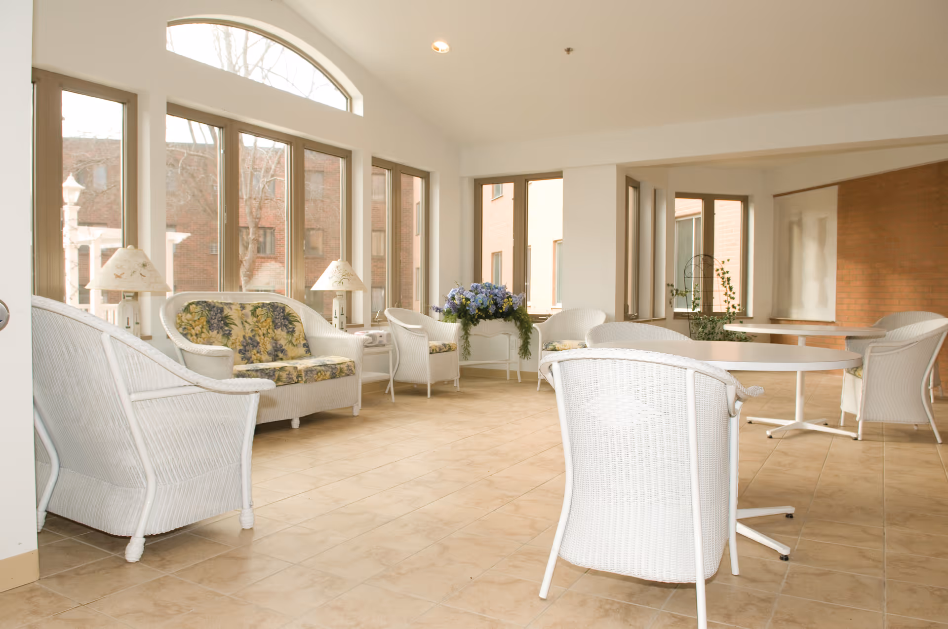 A bright and airy sitting area with white wicker furniture including chairs and a loveseat with floral cushions. There are two white tables and two lamps with floral lampshades. Large windows let in natural light and offer views of the outside buildings. A flower arrangement is placed on a small table near the windows.