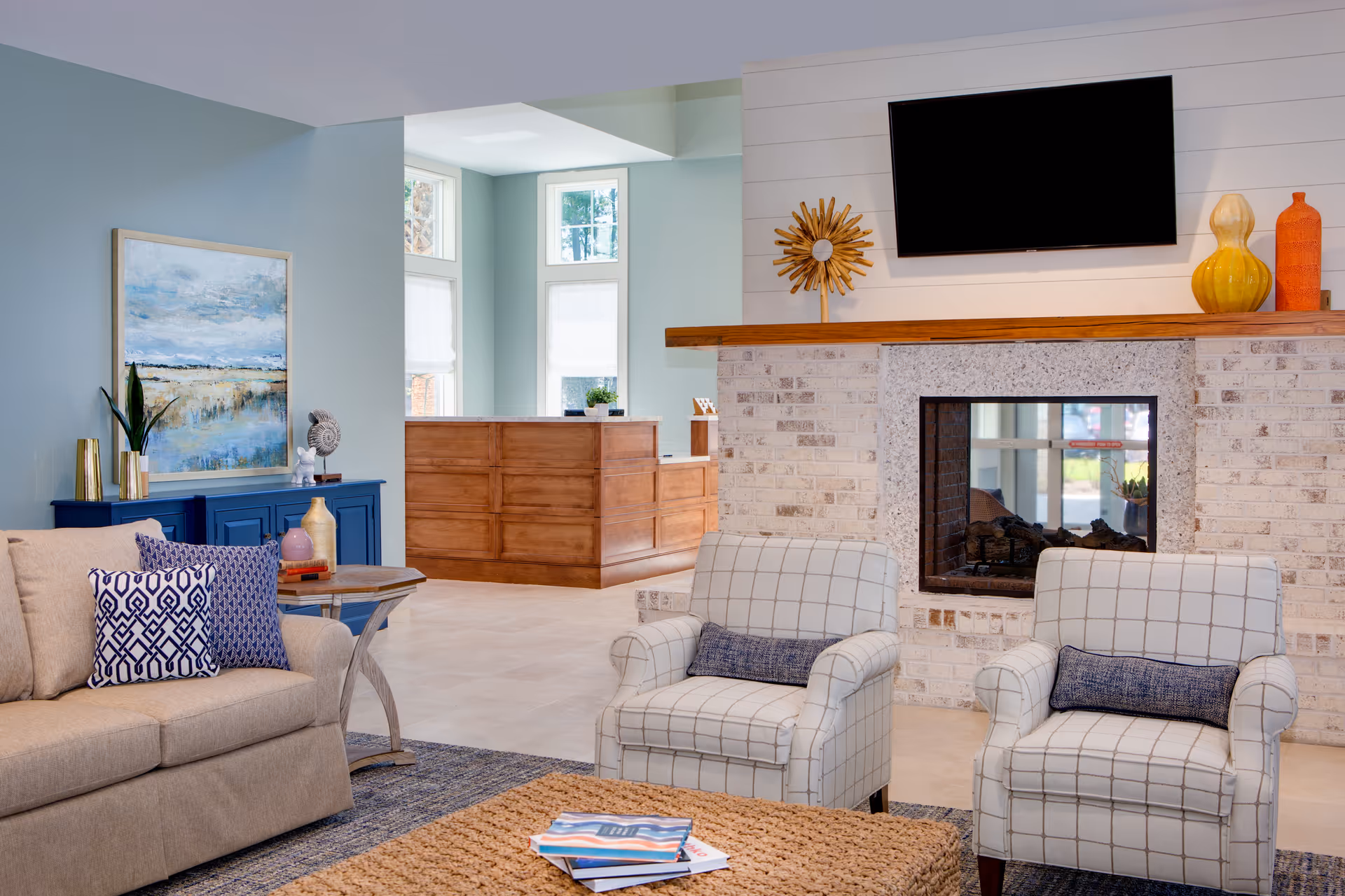 A cozy living room area with a beige sofa adorned with patterned blue and white pillows, two white armchairs with a grid pattern and blue lumbar pillows, a woven coffee table with magazines, and a brick fireplace with a wooden mantel. Above the mantel is a mounted flat-screen TV and decorative vases. In the background, there is a wooden reception desk and large windows letting in natural light.