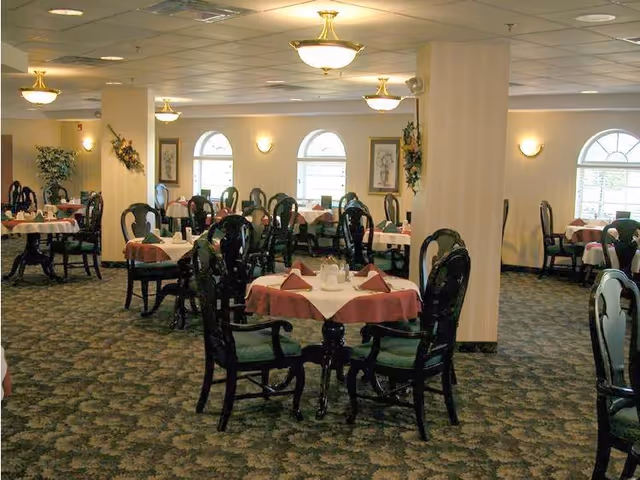 A dining room with multiple round tables covered with white and pink tablecloths, each set with folded napkins and glassware. The room has patterned carpet, arched windows, wall sconces, and ceiling lights, creating a warm and inviting atmosphere.