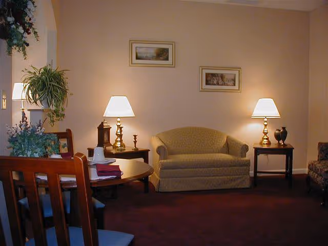A cozy living room area with a small beige loveseat centered between two wooden side tables, each with a brass lamp. There are framed pictures on the wall above the loveseat. To the left, a wooden dining table with chairs is partially visible, along with some green plants and decorative items.