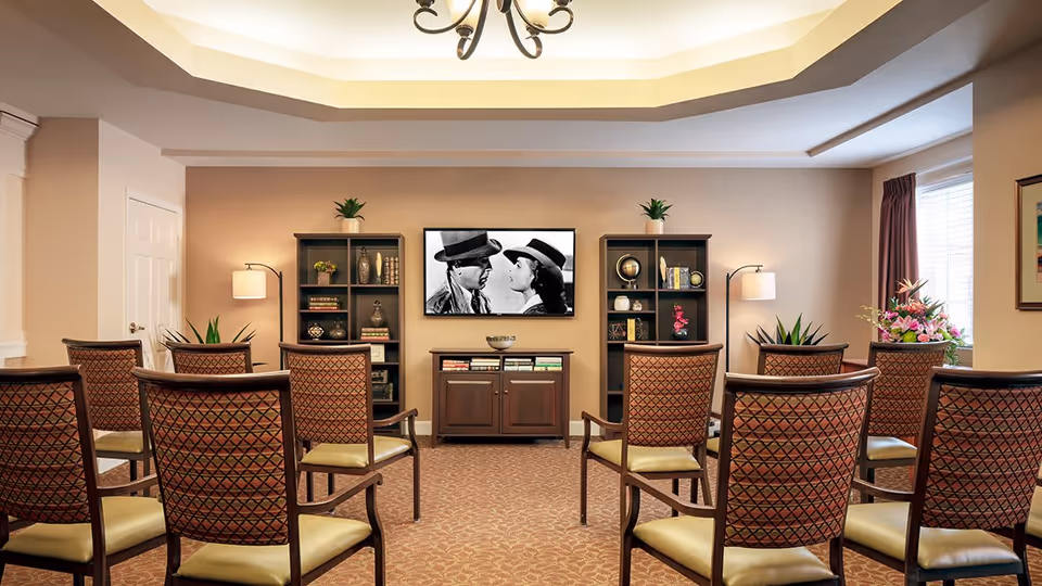 A cozy common area with rows of patterned chairs facing a wall-mounted TV displaying a black-and-white movie scene. The room features two dark wooden bookshelves with decorative items and books, a small cabinet below the TV, warm beige walls, a window with curtains on the right, and ceiling lighting with a chandelier.