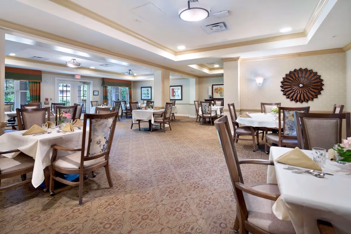 A spacious dining room in a senior living facility with multiple tables covered in white tablecloths, each set with beige napkins, glasses, and silverware. The room features wooden chairs with cushioned seats and backs, patterned carpet, soft lighting, and decorative wall art. Large windows and glass doors allow natural light to enter the room.