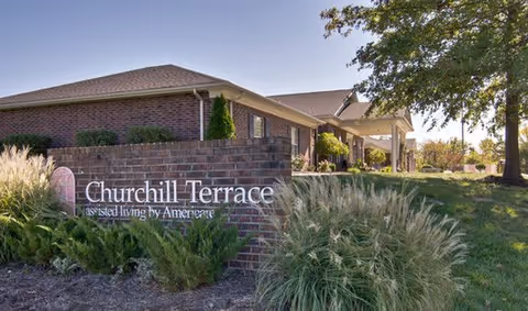 Exterior view of Churchill Terrace Senior Living facility showing a brick building with a sign in front that reads 'Churchill Terrace assisted living by Americare', surrounded by greenery and trees under a clear sky.