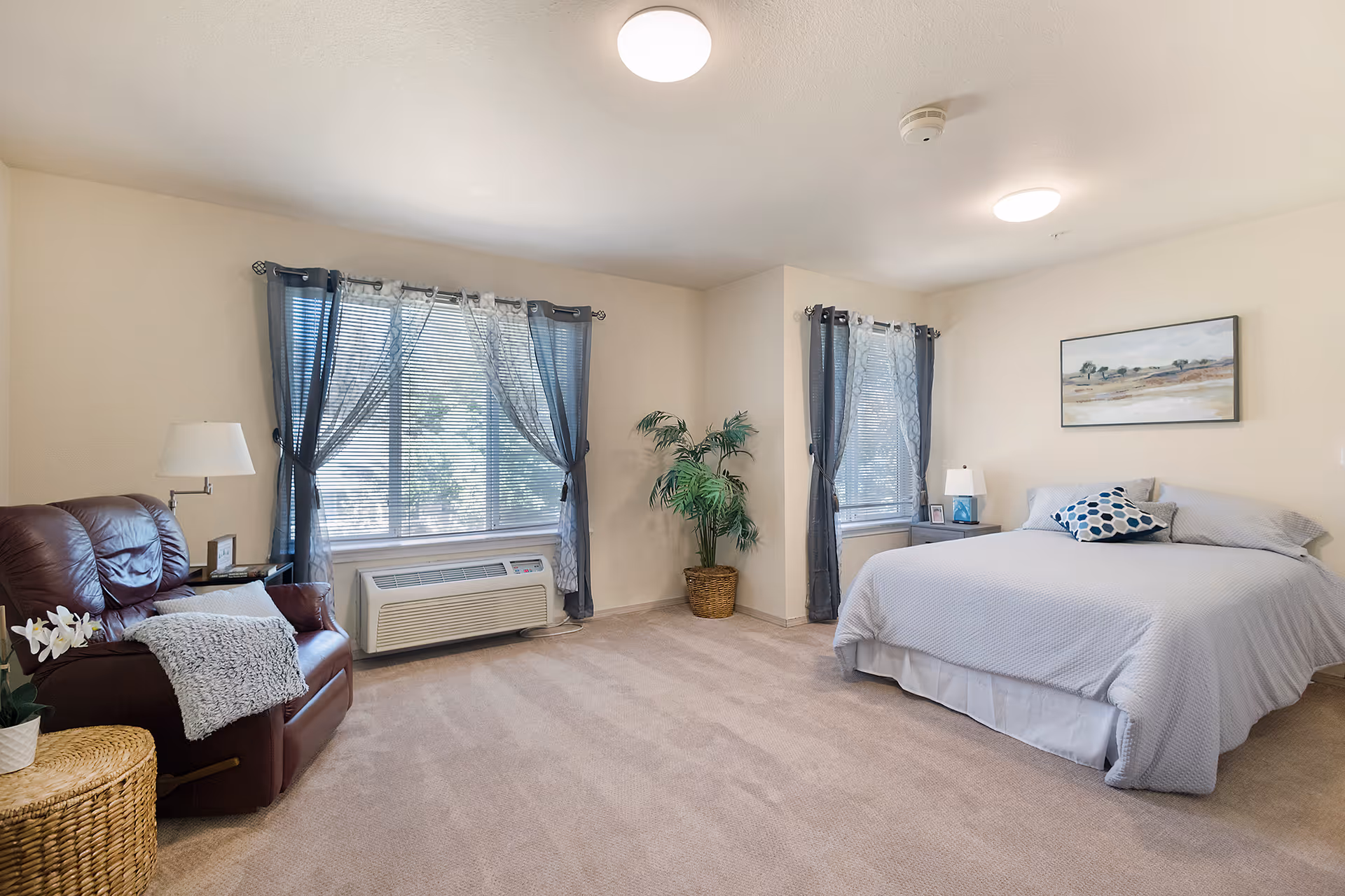 A spacious bedroom with beige walls and carpet. The room features a large bed with light gray bedding and a decorative pillow, two windows with sheer and dark curtains, a brown leather recliner chair with a gray throw blanket, a wicker side table with a white orchid plant, a potted plant in the corner, and two bedside tables with lamps. A landscape painting hangs above the bed.