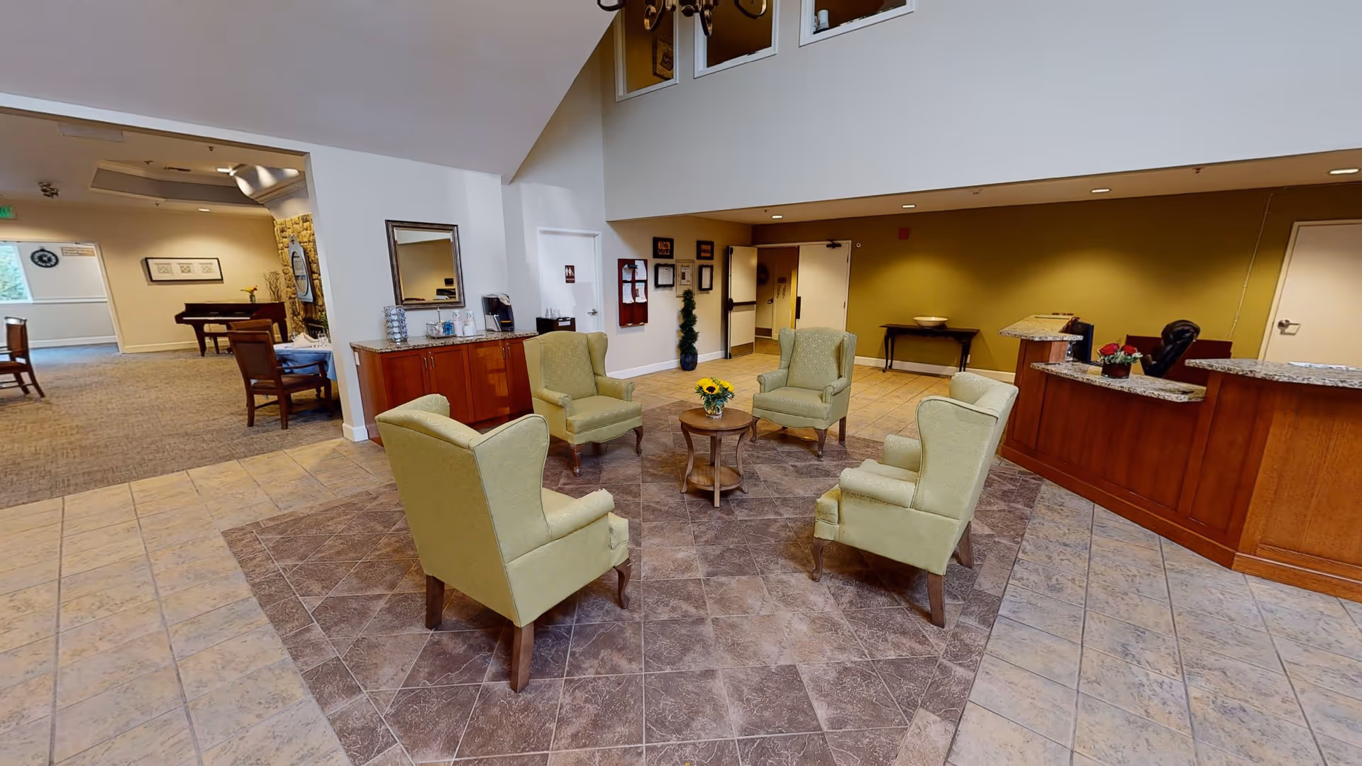 A spacious seating area in a retirement center with five light green armchairs arranged around a small round wooden table with a vase of sunflowers. The floor has a tiled pattern, and there is a wooden reception desk with granite countertops to the right. In the background, there is a hallway and another room with dining tables and chairs. The walls are painted in neutral tones with some framed pictures and a mirror on the left wall.