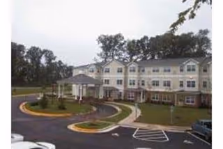Exterior view of a three-story senior living facility building with a covered entrance, surrounded by a driveway and landscaped greenery under a cloudy sky.