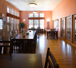 A spacious dining room with wooden floors and peach-colored walls. There are multiple dark wooden tables and chairs arranged neatly, with large windows letting in natural light. The windows have decorative valances, and there are two hanging light fixtures from the ceiling. French doors are visible on the right side of the room.