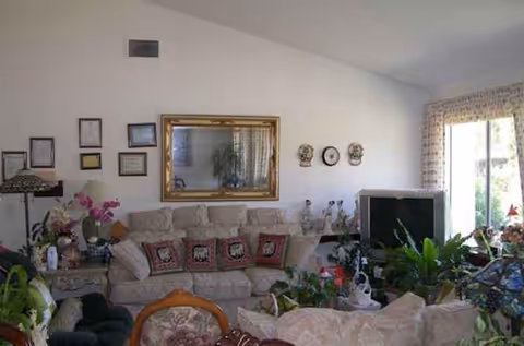 Cluttered senior living room with beige sofa, many plants, and a TV.