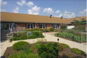 Outdoor view of a single-story brick building with a sloped roof under a partly cloudy sky. The foreground features a landscaped garden with various green shrubs and plants, along with paved walkways and metal handrails leading to the building entrance.