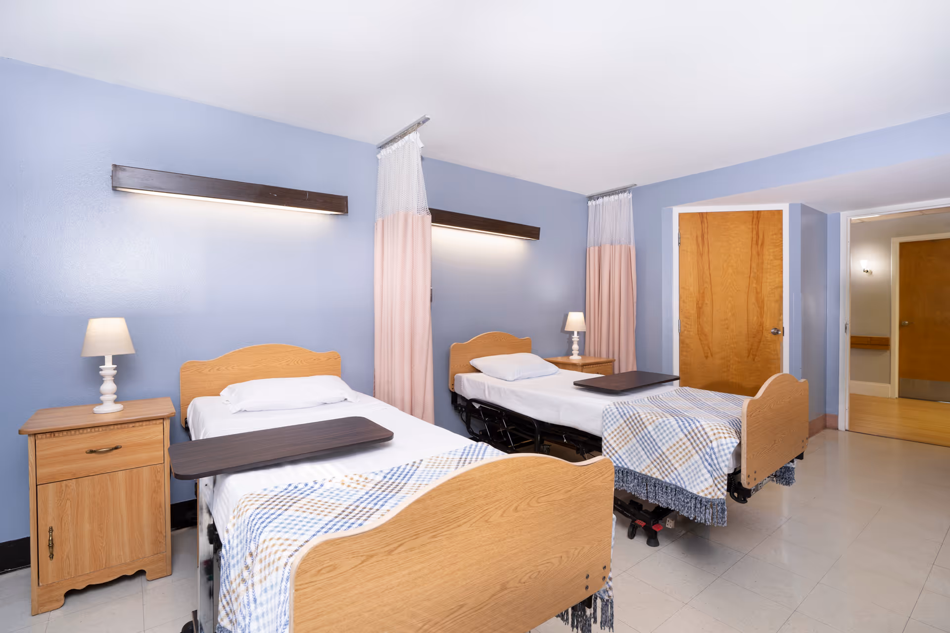 A room with two hospital-style beds each with a wooden headboard and footboard, white sheets, a pillow, and a plaid blanket. Each bed has a small overbed table. There are two wooden nightstands with lamps on either side of the beds. The walls are painted light blue and have two long horizontal lights above the beds. Pink privacy curtains hang between the beds and near the door. The floor is tiled, and there are two wooden doors visible in the background.