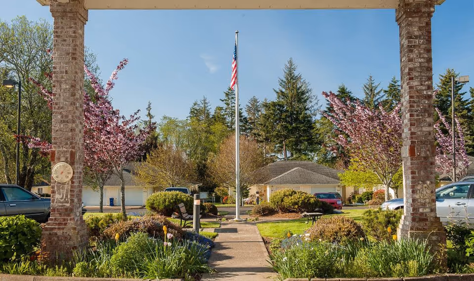 View of a landscaped outdoor area at Lakeview Senior Living with a pathway leading to an American flagpole surrounded by bushes, flowering trees, and parked cars. The scene is framed by two brick pillars under a covered structure.