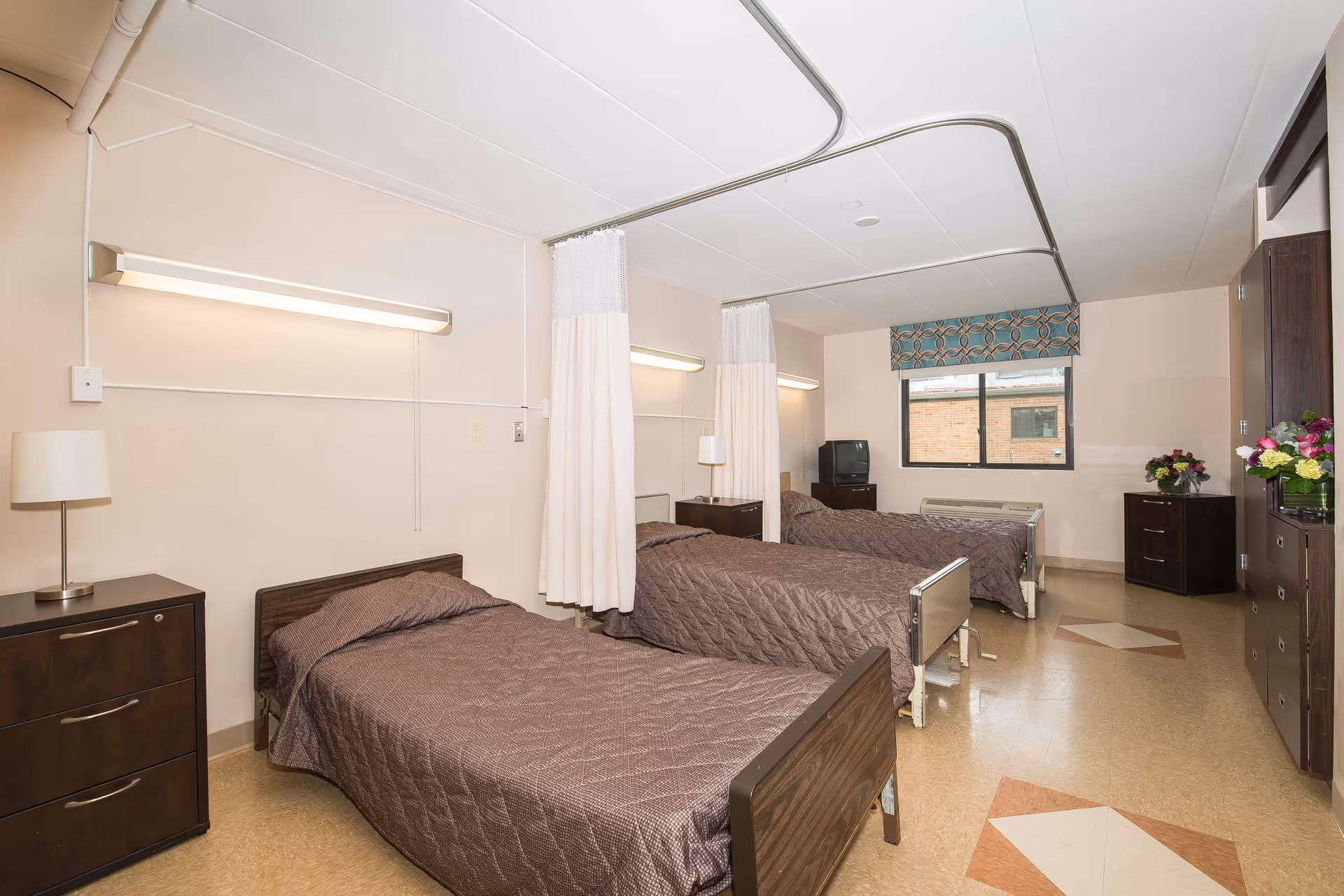 A room with three single beds arranged in a row, each with brown quilted bedspreads. There are privacy curtains hanging from ceiling tracks between the beds. The room has beige walls, a window with a patterned valance, and several dark wood nightstands and dressers. A small television is placed on one of the dressers near the window.