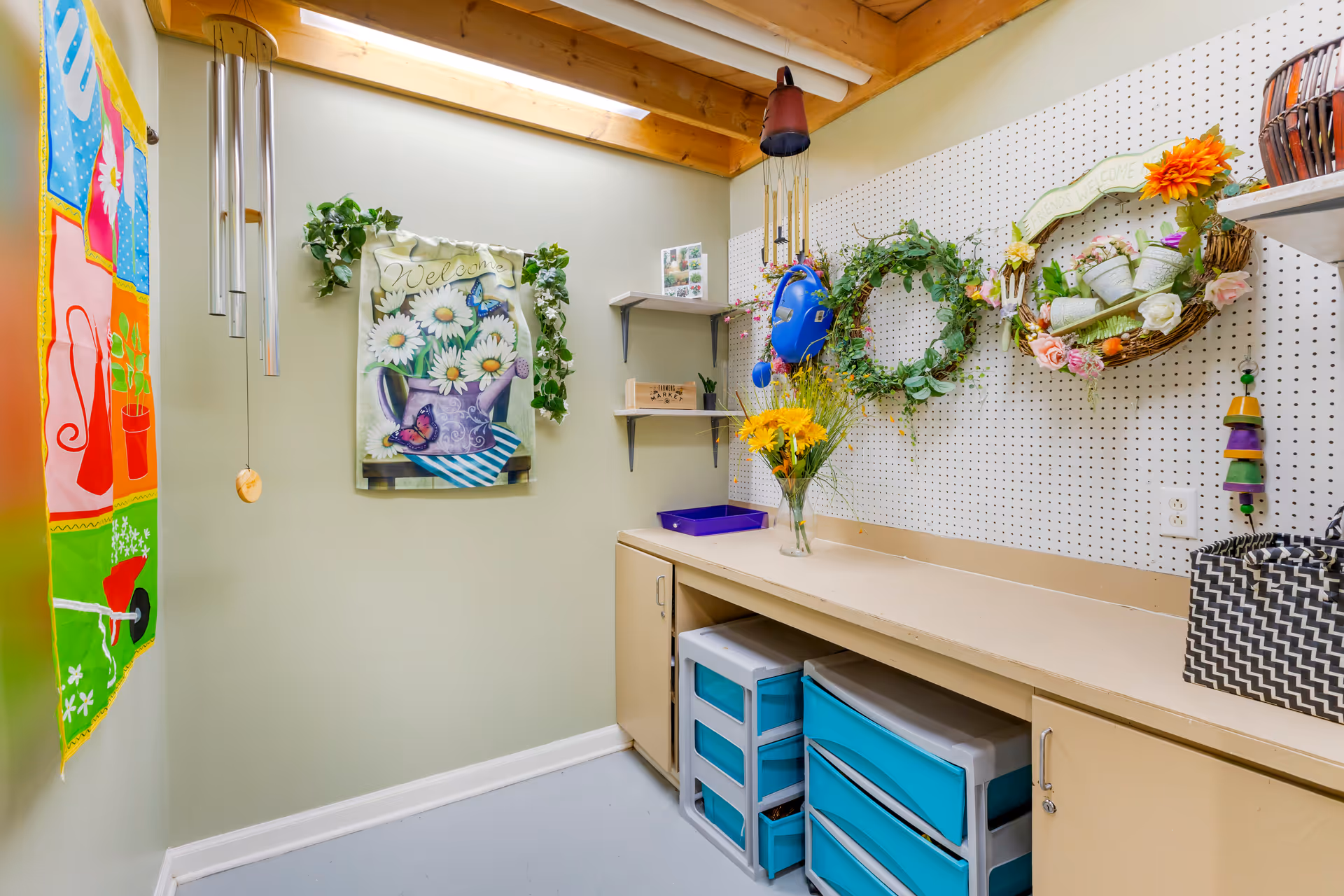 Small craft room with a pegboard wall displaying wreaths and decorations, a countertop with storage drawers and a vase of flowers.