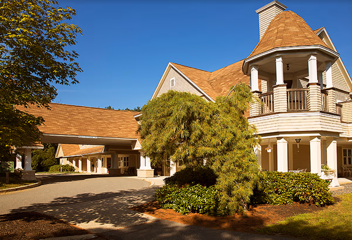 Exterior view of a senior living facility building with beige siding and a brown shingled roof under a clear blue sky. The building features a covered entrance driveway and a small tower-like structure with a balcony. There are trees and shrubs around the building.