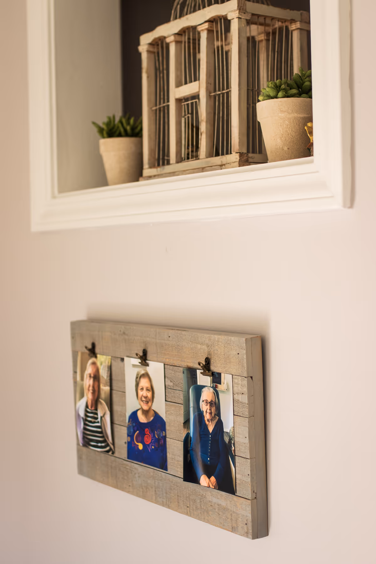 A wooden photo frame mounted on a beige wall displaying three photos of smiling elderly women. Above the frame, there is a white-framed window-like opening with two small potted plants and a decorative wooden birdcage inside.