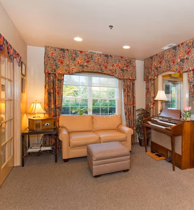 A cozy living room with a beige leather sofa and matching ottoman in front of a large window with floral curtains. To the right, there is a wooden piano with a lamp and a small plant on top. On the left side, a small table with a vintage radio and a lamp is placed next to a glass-paneled door. The room is carpeted and well-lit with recessed ceiling lights.