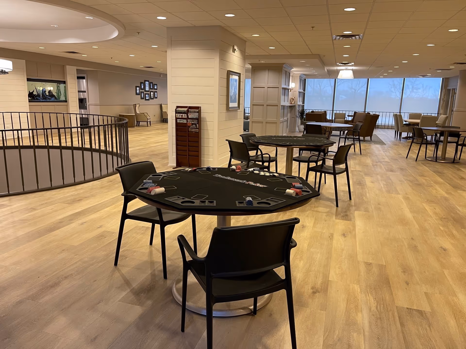 A spacious, well-lit common area in a retirement community featuring multiple round tables with black chairs. Two tables in the foreground are set up for poker with chips and cards. The room has light wood flooring, large windows in the background, and comfortable seating areas with armchairs. The walls are decorated with framed pictures and a magazine rack is visible near a column.