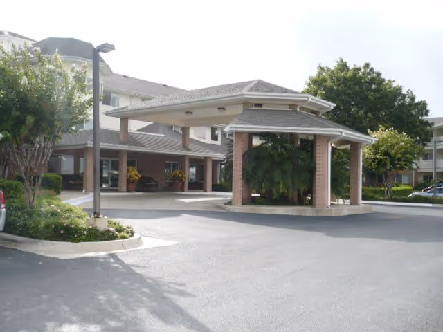 Entrance area of a senior living facility with a covered drop-off zone supported by brick columns, surrounded by trees and shrubs, with a multi-story building in the background.