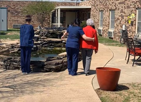 An outdoor courtyard area at a senior living facility with a stone water feature. Two women in navy blue uniforms are assisting an elderly woman wearing a red sweater as they walk along a curved concrete path. Another woman in a navy blue uniform stands nearby looking at the water feature. The building's brick exterior and windows are visible in the background.