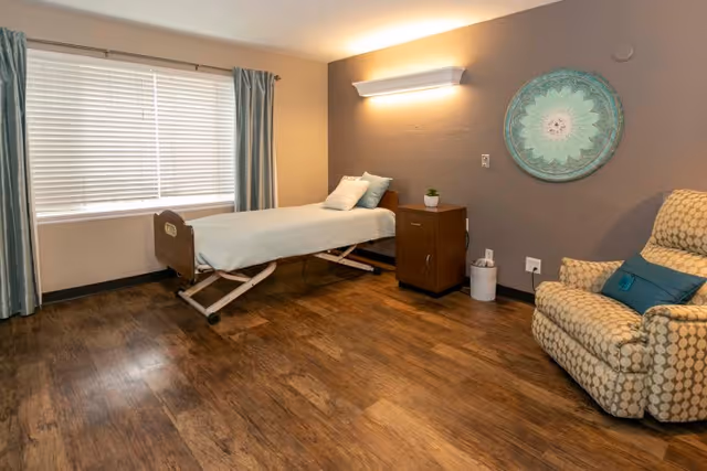 A simple, clean room with a single adjustable bed positioned near a large window with closed blinds and light blue curtains. Next to the bed is a wooden nightstand with a small plant on top. On the right side of the room is a patterned armchair with a blue cushion. The walls are painted in neutral tones, and a decorative round wall art piece hangs above the nightstand. The floor is covered with wood-like vinyl flooring.