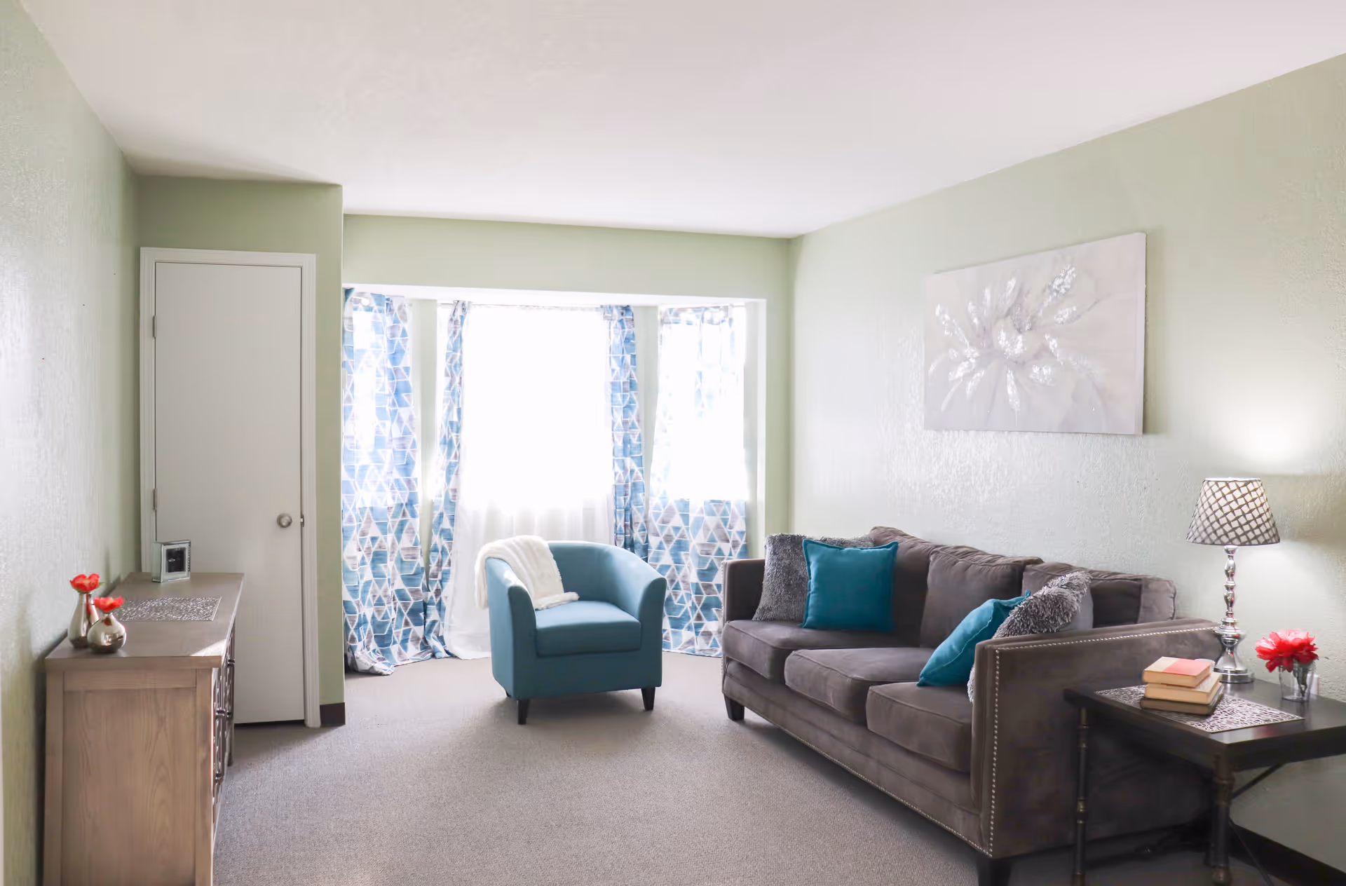 Bright living room with a gray sofa and teal armchair by a bay window with patterned curtains, side tables and decor.