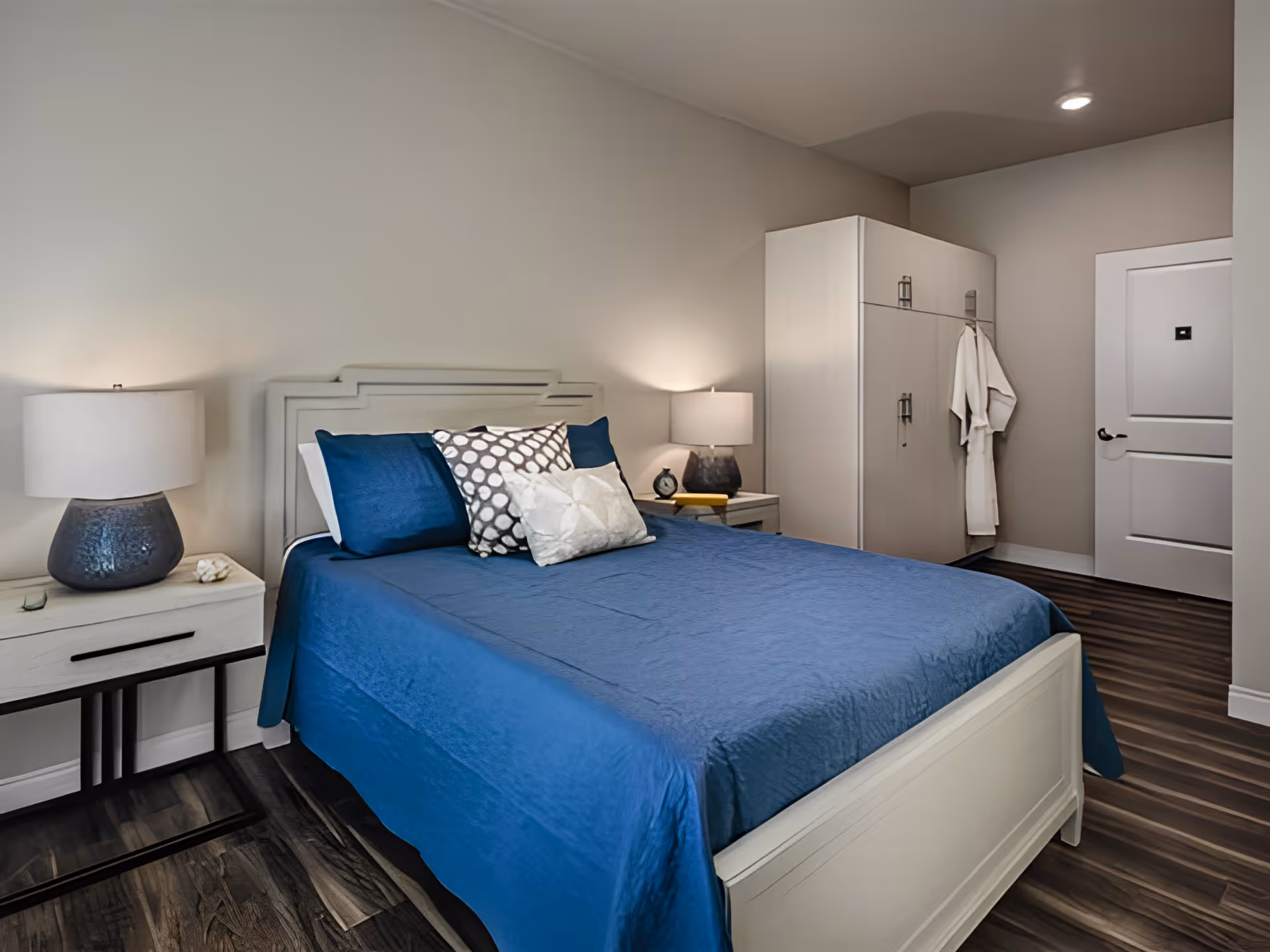 A tidy bedroom with a blue-covered bed, two nightstands with lamps, a wardrobe with a hanging robe, and a closed door.