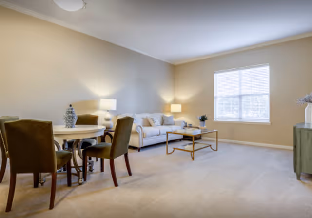 Bright furnished living room with a sofa, coffee table, round dining table with four chairs, lamps, and a window.