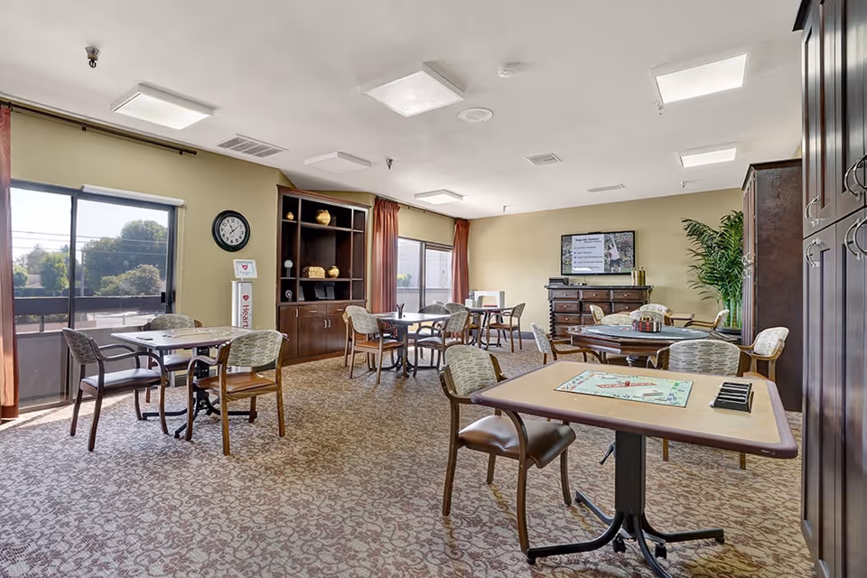 A well-lit common area with multiple tables and chairs arranged for socializing or activities. The room has large windows with curtains, a carpeted floor, a wall clock, a wooden cabinet with decorative items, a TV mounted on the wall displaying information, and a large potted plant in the corner. One table has a board game set up on it.