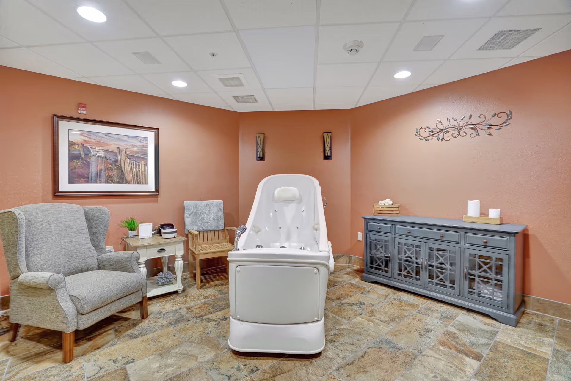 A cozy room with a large white therapeutic bathtub in the center, surrounded by a gray upholstered armchair, a wooden chair, a small side table with a plant and a device on it, and a blue cabinet with decorative items on top. The walls are painted a warm terracotta color, and there is a framed landscape painting and wall sconces for lighting.