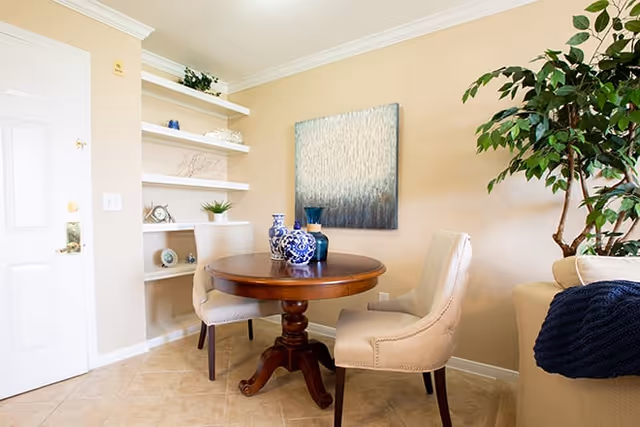 Small dining nook with a round wooden table, two upholstered chairs, decorative blue vases, wall art, built-in shelves, and a potted plant.