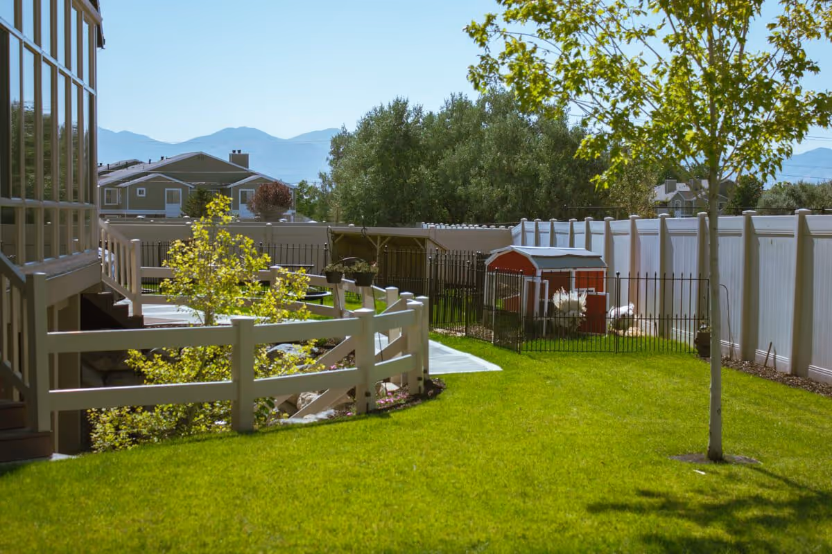 A sunny outdoor garden area at Assisted Living Of Draper featuring a well-maintained green lawn, a white fence, a small tree, and a fenced enclosure with a red and white chicken coop. In the background, there are trees, residential houses, and mountains under a clear blue sky.