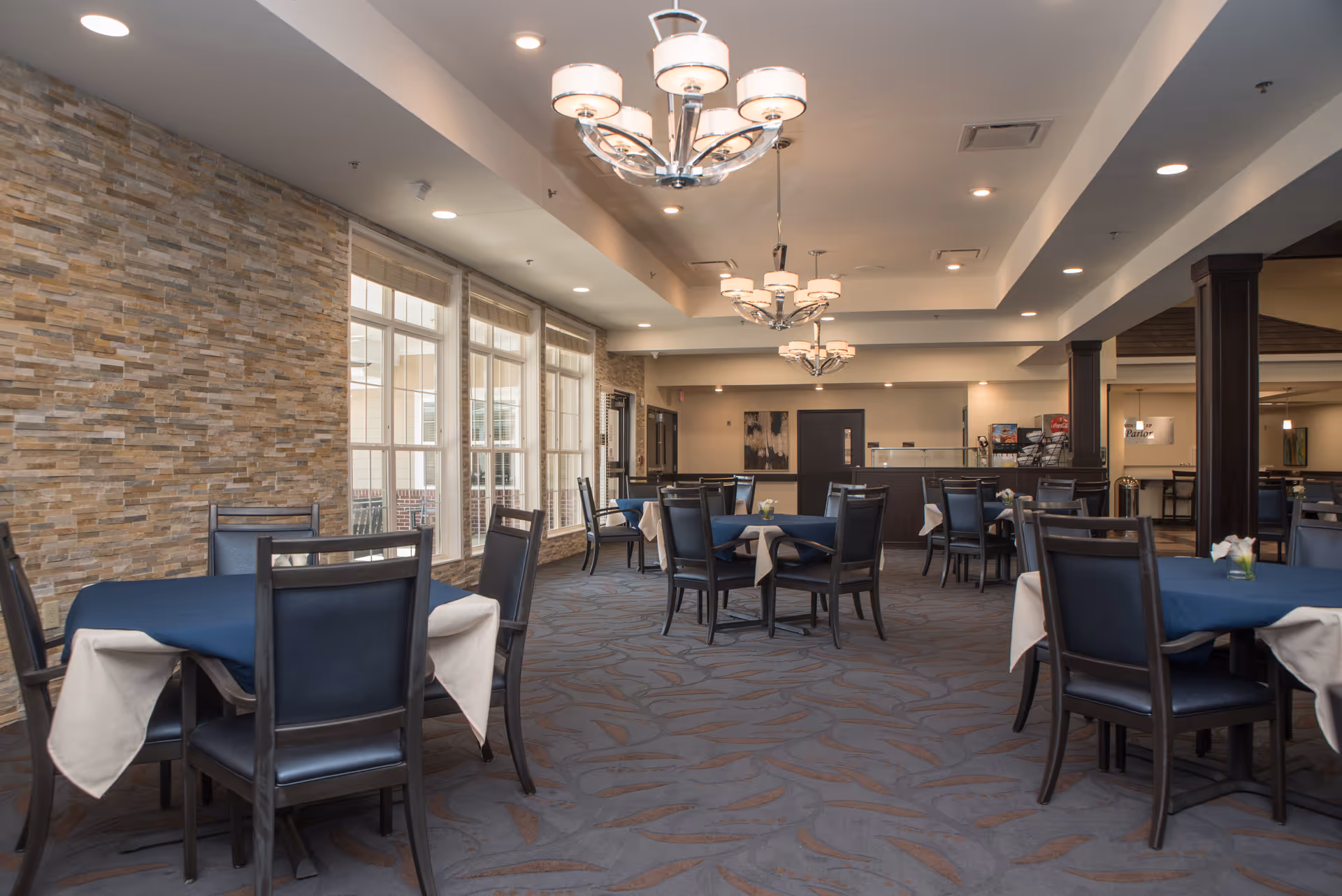 Spacious dining room with round tables draped in blue tablecloths, dark chairs, chandeliers, and large windows along a stone accent wall.