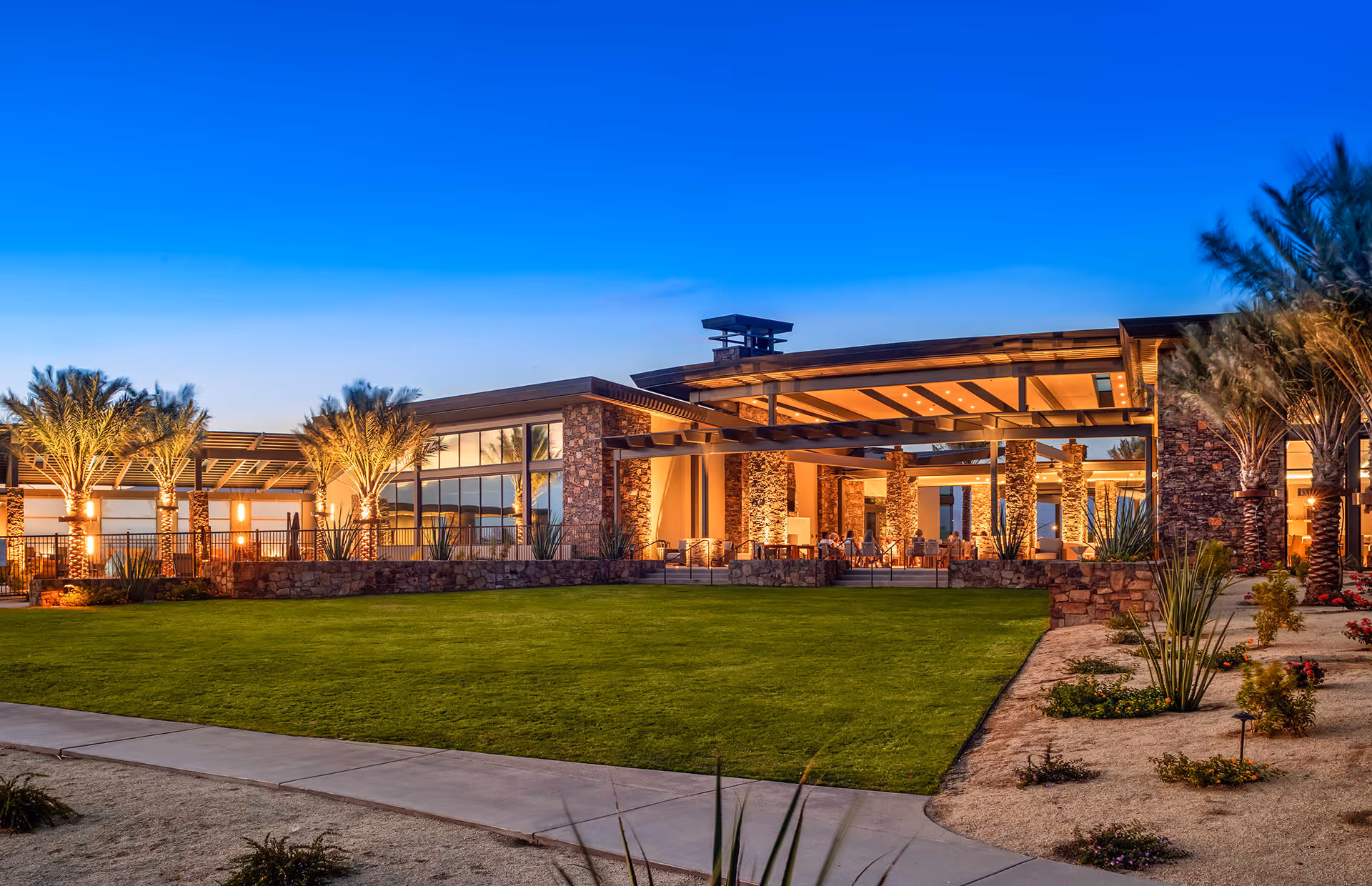 Exterior view of a modern retirement community building at dusk with large glass windows, stone pillars, palm trees, and well-maintained green lawn and landscaping.