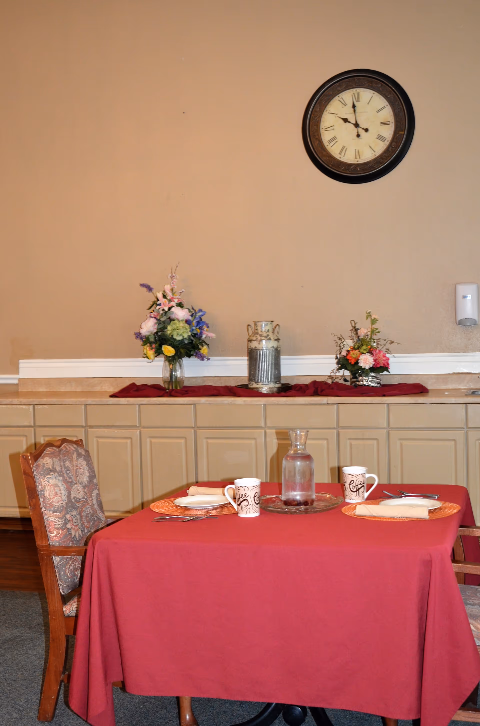 A dining table covered with a red tablecloth set for two with plates, napkins, coffee mugs, and a glass water pitcher. Behind the table is a counter with floral arrangements and a decorative vase. A round wall clock shows the time as 11:55. The wall is beige, and there is a hand sanitizer dispenser mounted on the right side.
