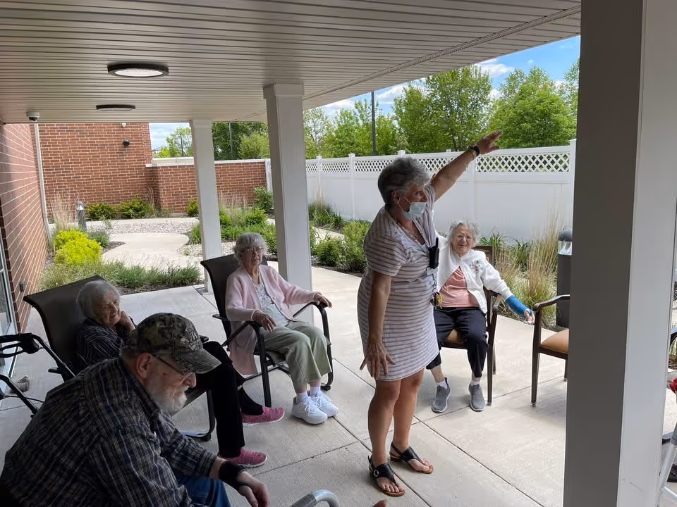 A group of elderly people sitting on chairs under a covered patio area outside, with one woman standing and raising her arm, wearing a face mask. The background shows a white fence, green trees, and a brick wall.