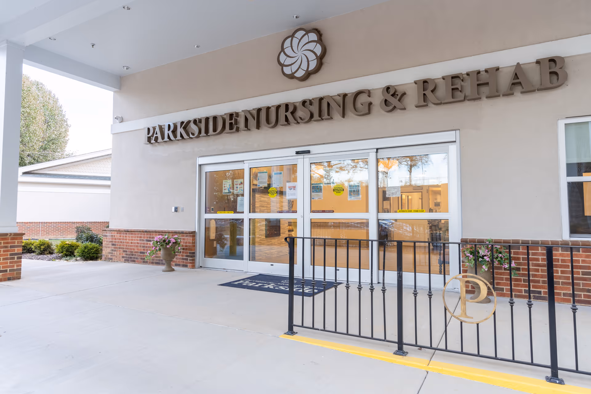 Entrance to Parkside Nursing & Rehab facility with automatic sliding glass doors, a covered walkway, brick and beige exterior walls, potted plants, and a black metal railing with a gold circular emblem.