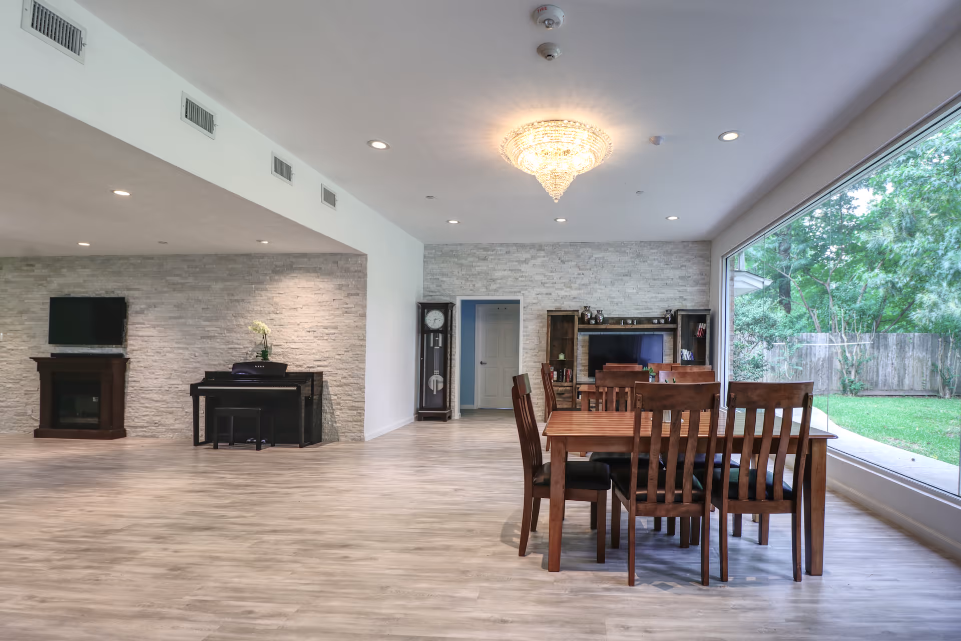 Spacious communal interior with a wooden dining table and chairs, large window overlooking a lawn, piano, TV and fireplace against a stone accent wall.