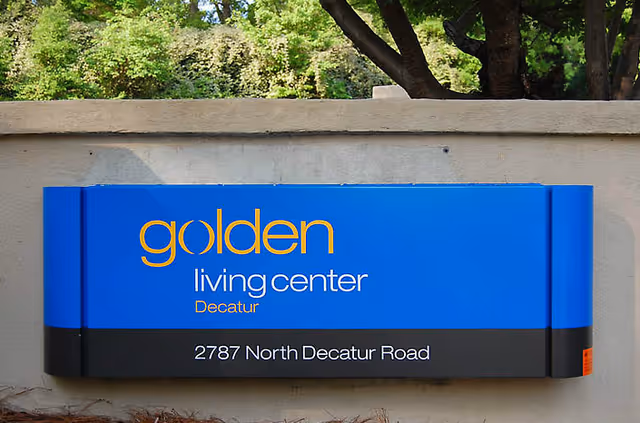 Blue outdoor sign on a concrete wall reading "golden living center Decatur" with the address 2787 North Decatur Road and trees behind it.